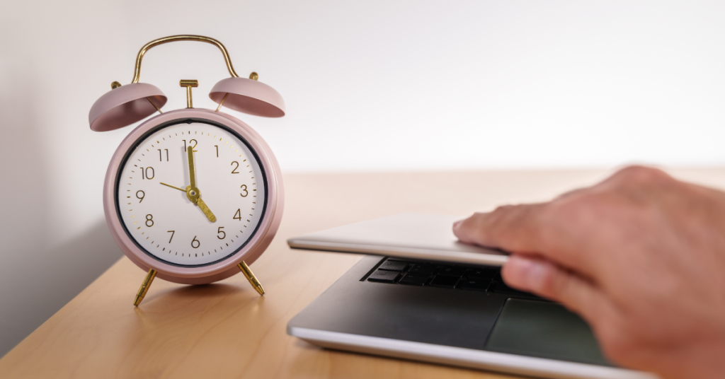 A hand closing a laptop that is next to a pink desk clock.